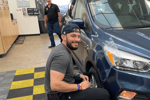 Student smiling looking at the camera while he's sitting next to the PPF work he's doing to a headlight on a car.