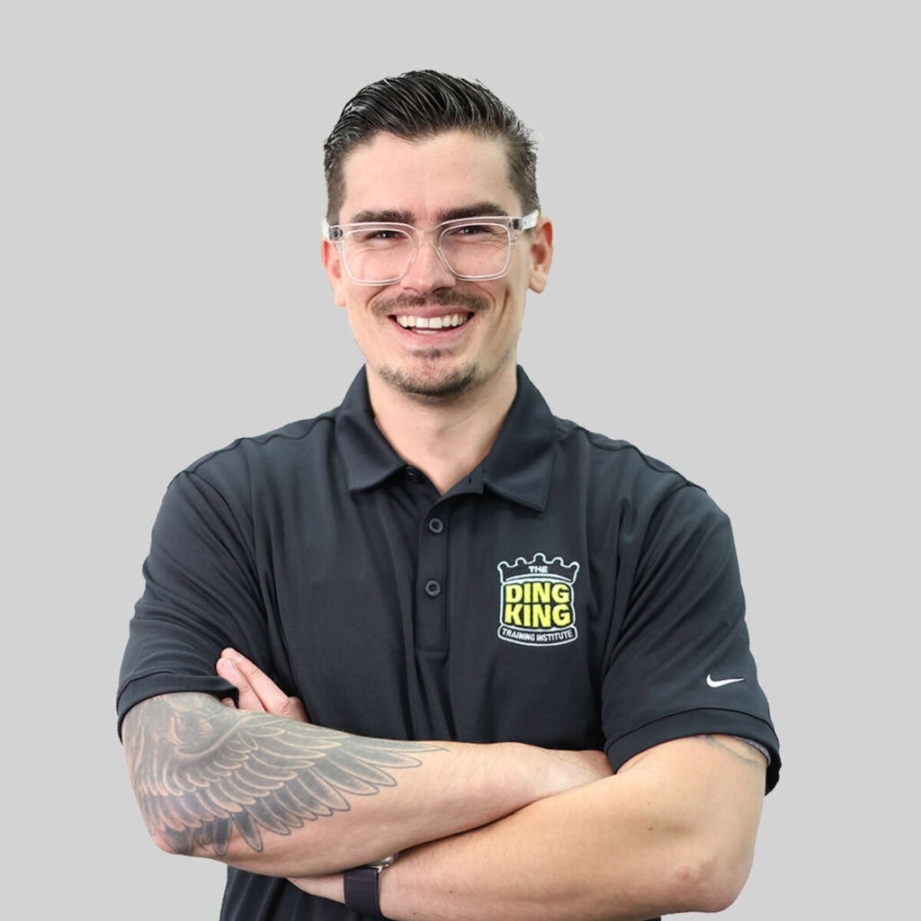 Man with a beard and glasses, wearing a black team polo shirt with "ding king" logo, smiling with arms crossed, showcasing a tattoo on his left arm, against a gray background.