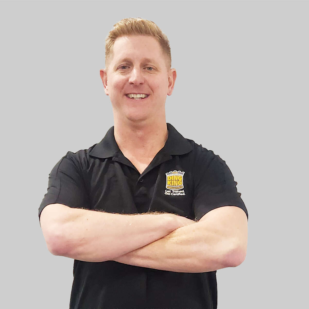 A smiling man with blond hair, wearing a black team polo shirt with a Burger King logo, standing with his arms crossed against a gray background.