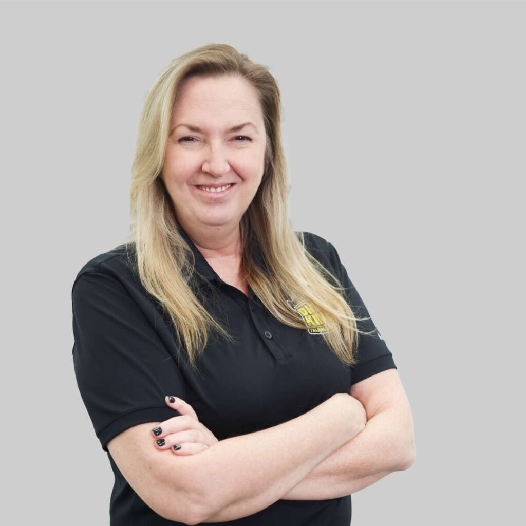 A smiling woman with blond hair, wearing a black polo shirt with a "meet the team" logo, stands with her arms crossed against a gray background.