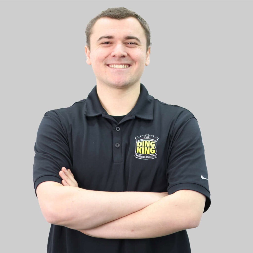 A man in a black team polo shirt with a "ding king" logo, smiling and crossing his arms, stands against a light gray background.