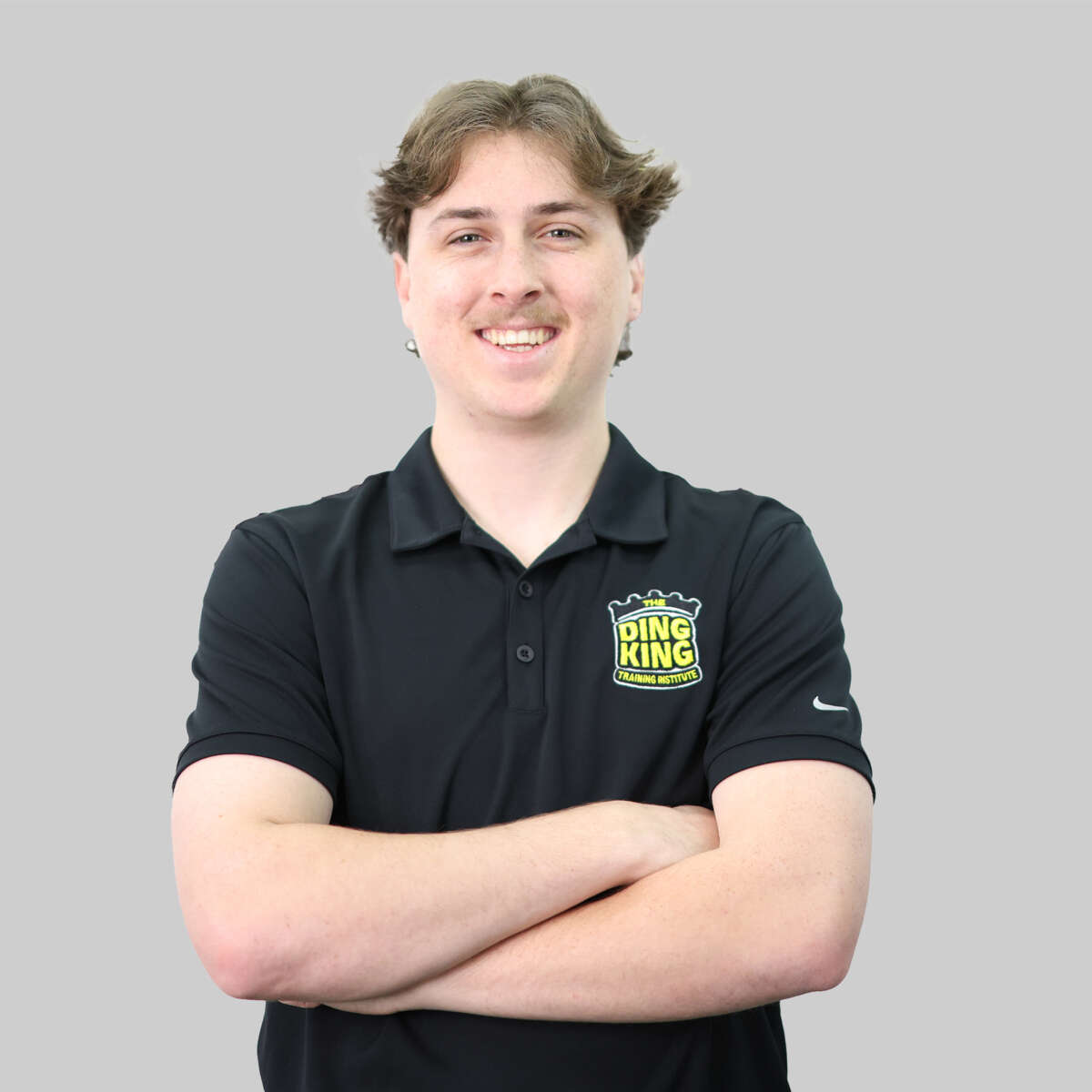 A smiling man with medium length hair, wearing a black polo shirt with a "king king" logo, is part of our meet the team series. He stands with crossed arms against a gray background.