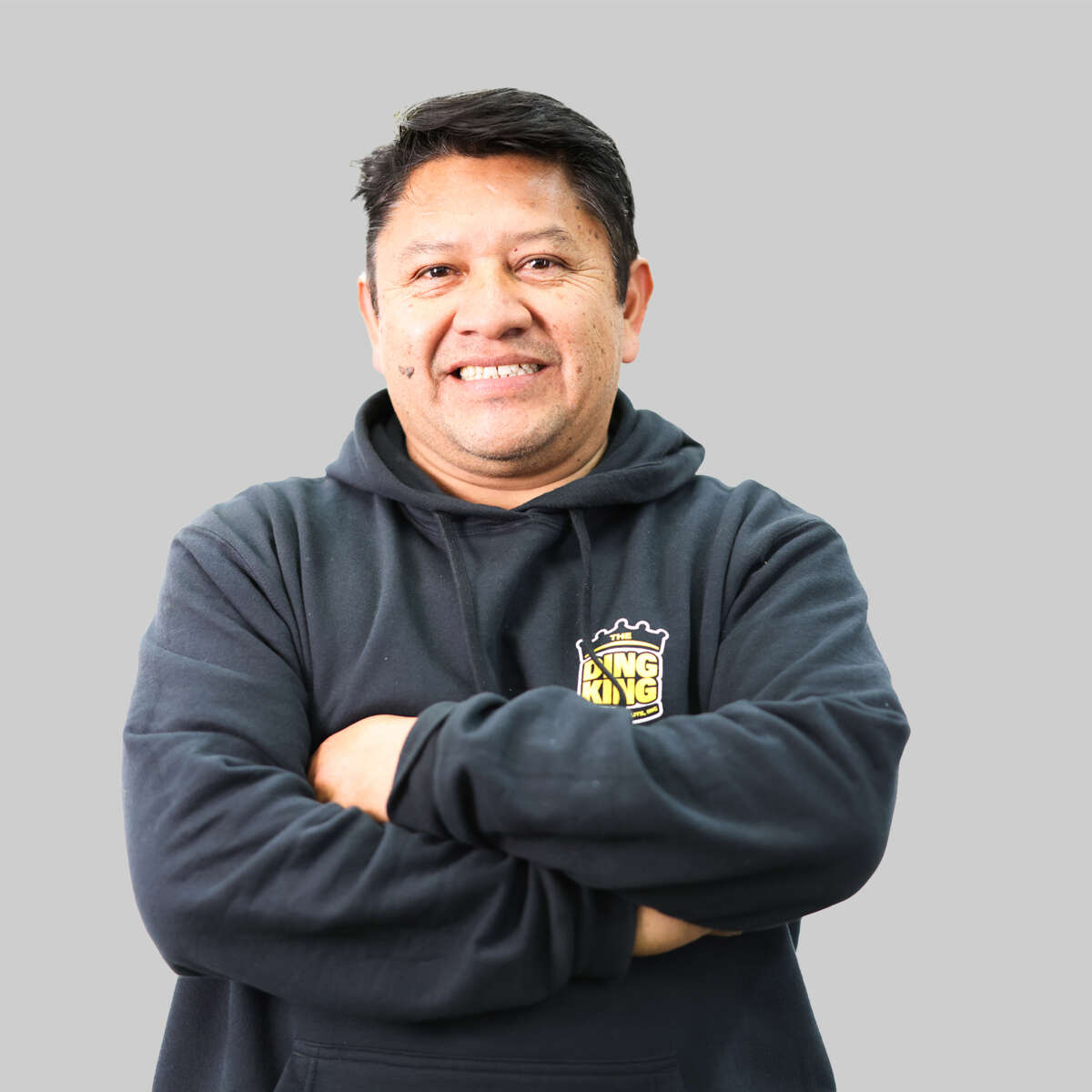 Middle aged man smiling at the camera, arms crossed, wearing a dark hoodie with a team logo, standing against a light gray background.