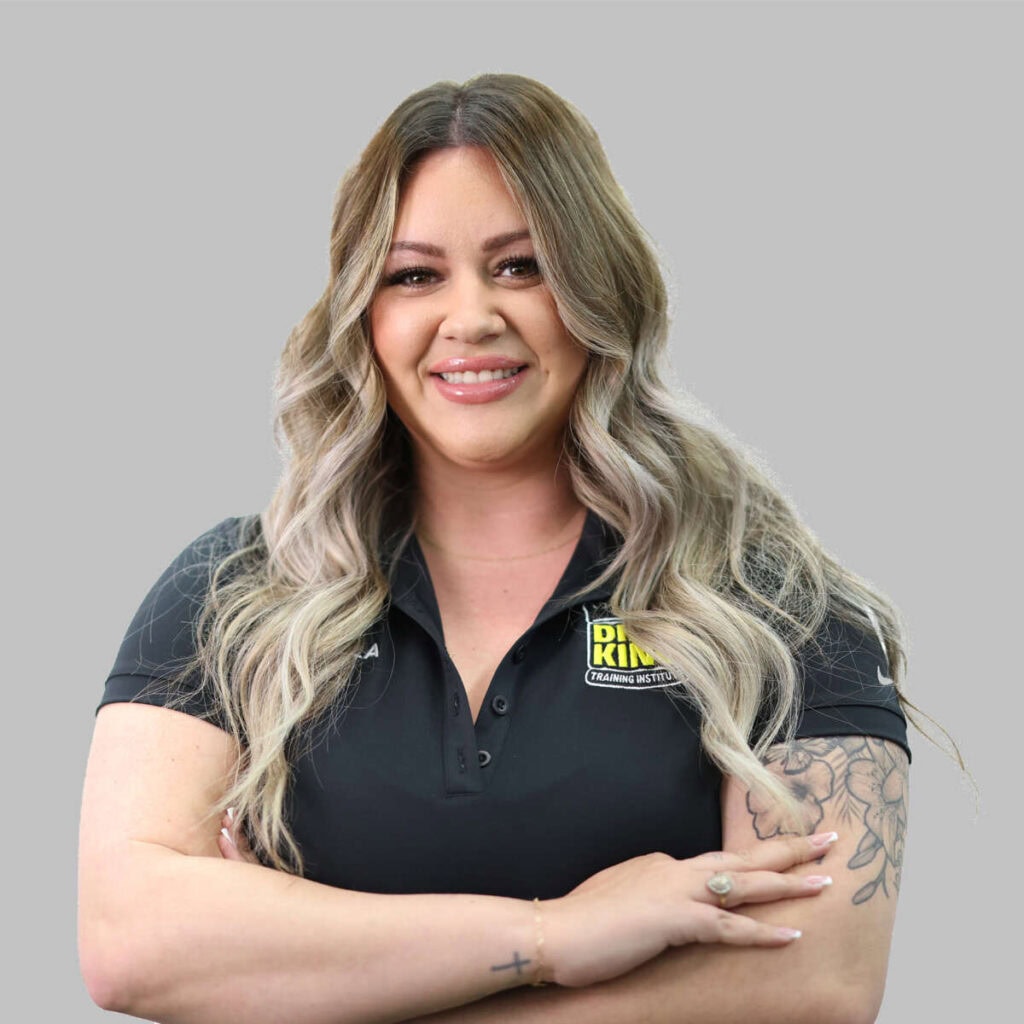 Woman in a black team polo shirt with a "dr. king" badge smiles, standing with crossed arms against a gray background. She has a tattoo on her left arm.