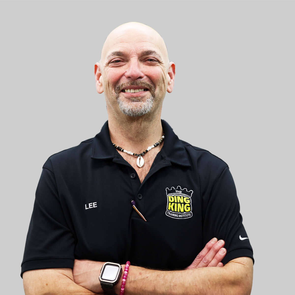 A smiling bald man wearing a black team polo shirt with "ding king" logo, arms crossed, wearing a watch and bracelet, against a grey background.