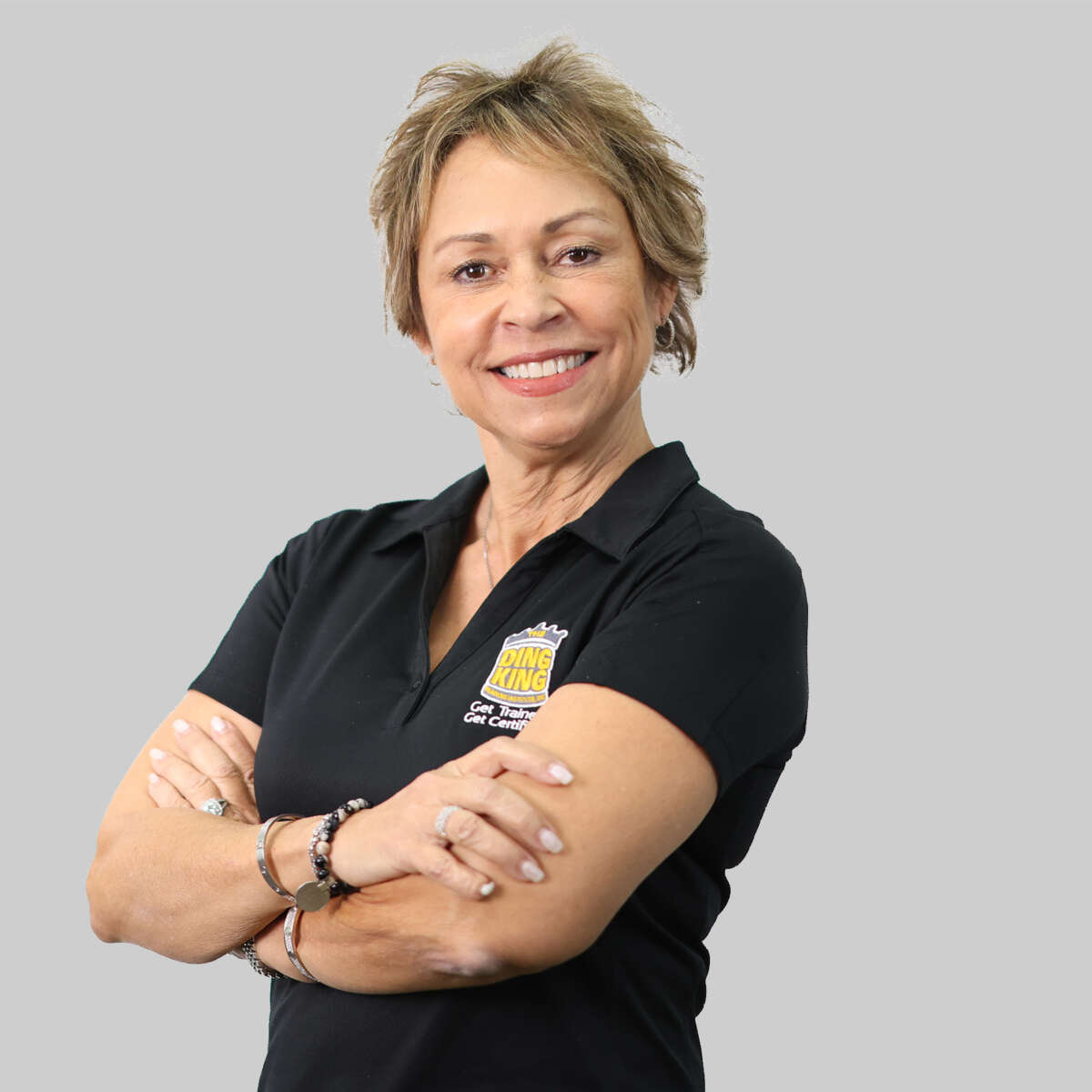 Mature woman with short hair, smiling, arms crossed, wearing a black polo shirt with a "King Cleaners" logo, standing against a gray background as part of our team members feature.