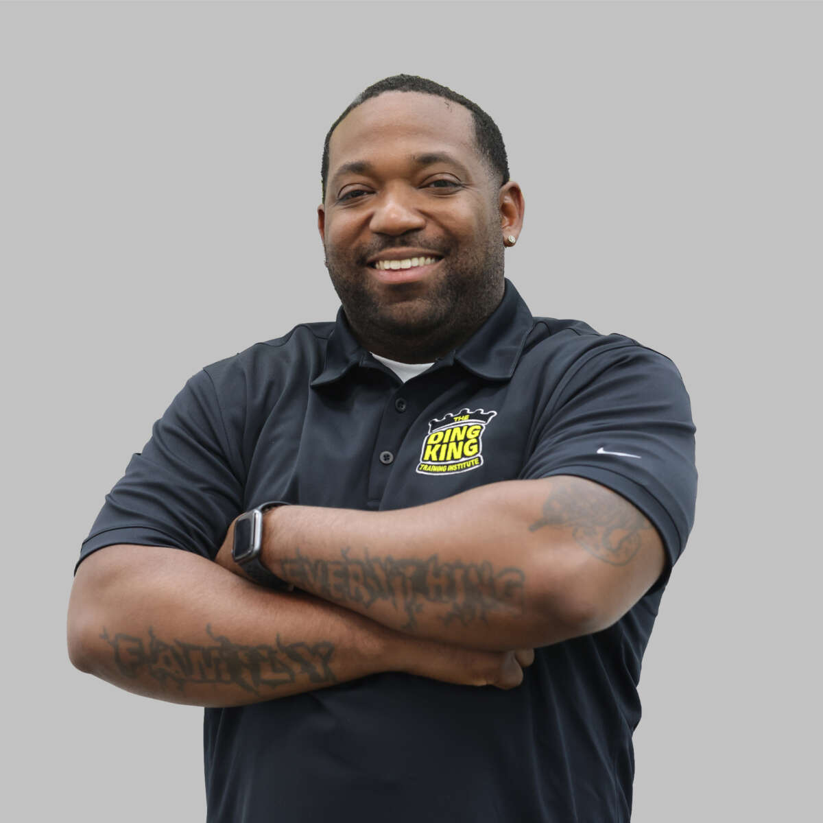 Smiling man with crossed arms wearing a "team ring king" polo shirt, tattoos visible on forearms, standing against a gray background.