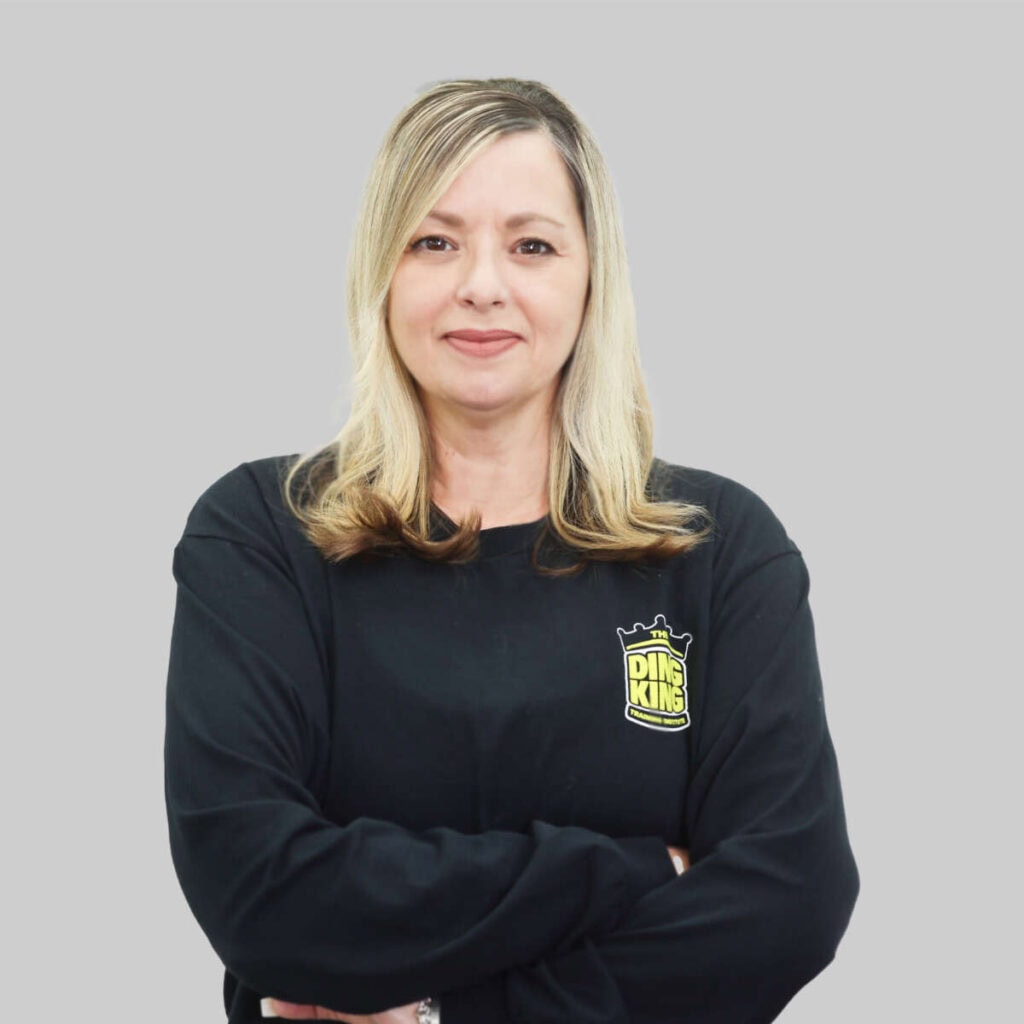 A middle aged woman with blonde hair stands with arms crossed, wearing a black sweatshirt with a "meet the team" logo, against a gray background.