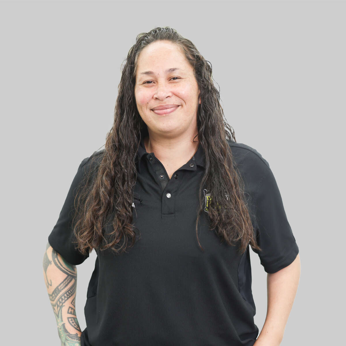 Meet our team: Woman with long curly hair, smiling, wearing a black polo shirt and displaying visible tattoos on her left arm, representing our company team against a gray background.