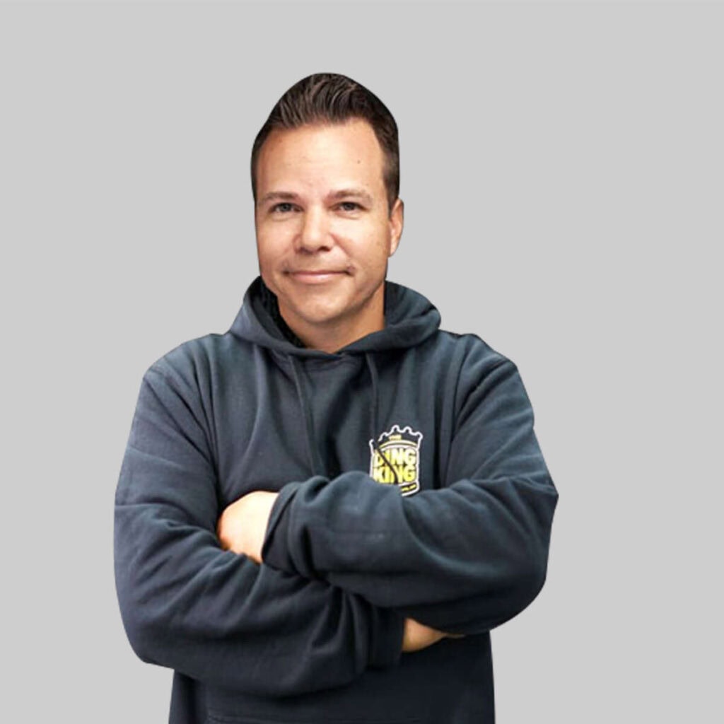 Man in a grey team hoodie with arms crossed, smiling slightly, standing against a light gray background.