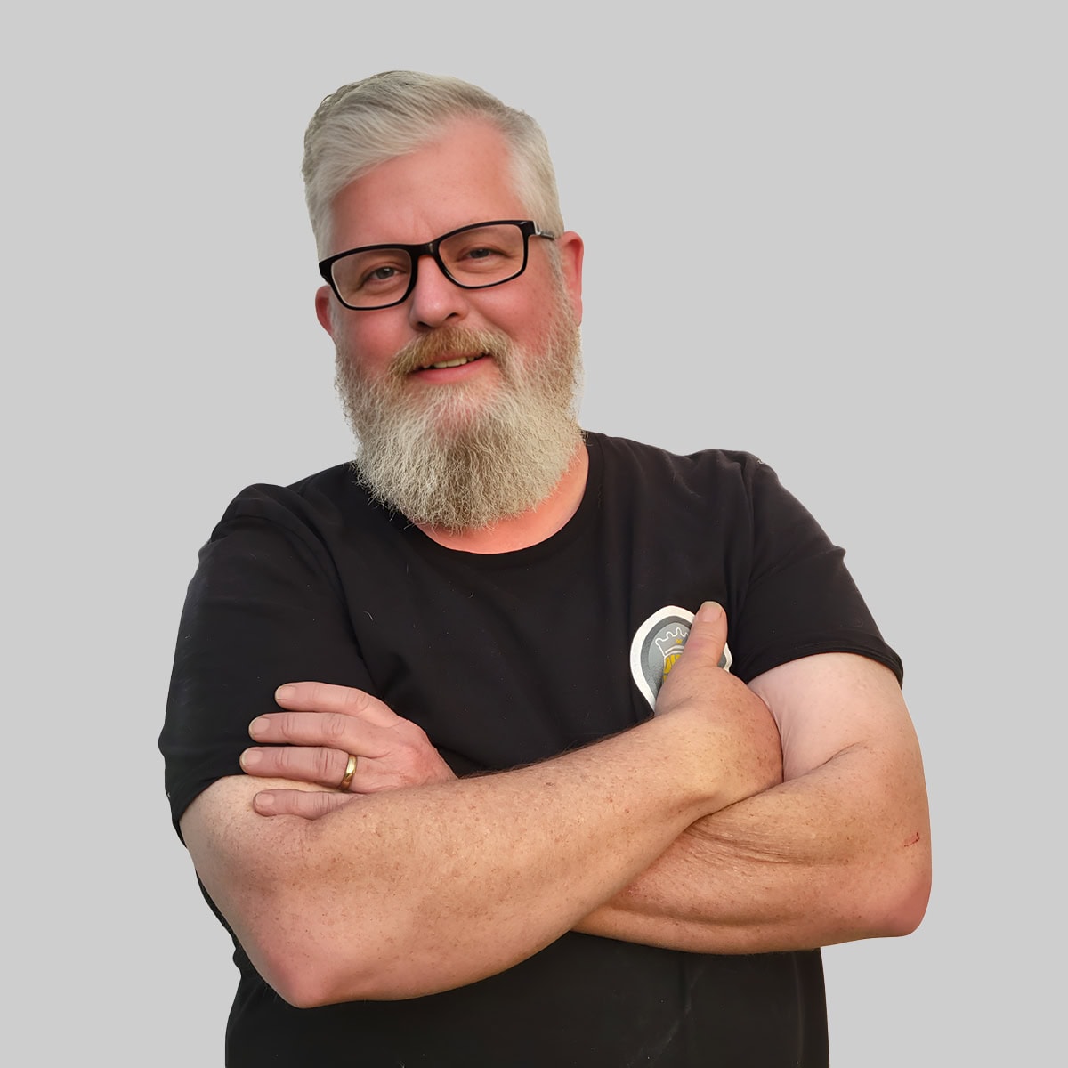 A smiling man with a beard wearing glasses and a black t shirt, standing with his arms crossed against a light gray background, ready for team introduction.