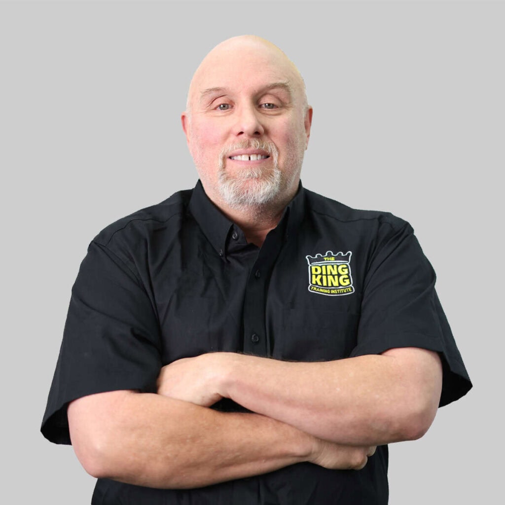 A bald man with a beard, wearing a black shirt with a "ring king" team logo, smiling and standing with his arms crossed against a gray background.