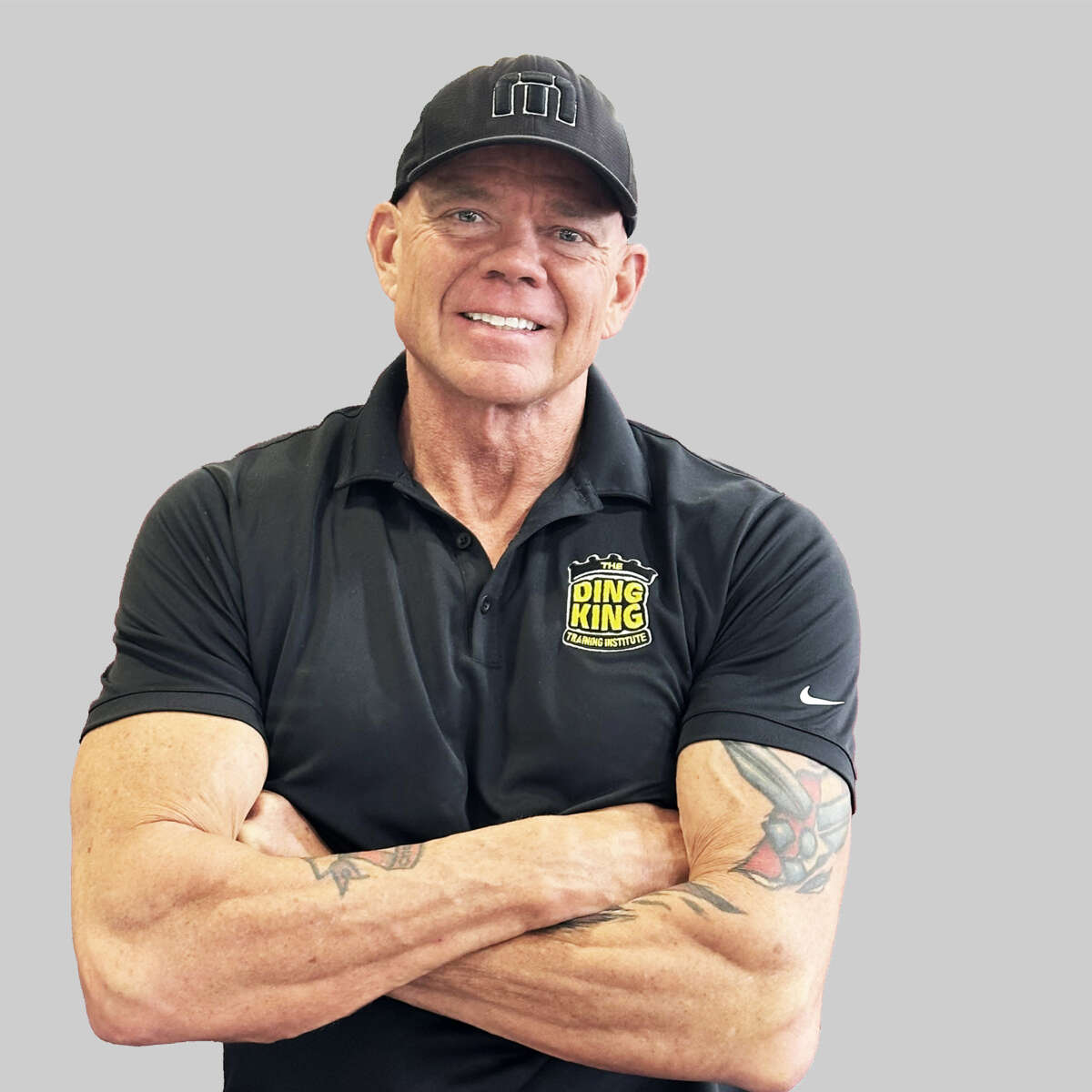 A fit middle aged man wearing a black team polo shirt with a "king kong nutrition" logo, smiling with arms crossed, sporting a tattoo on his left arm.