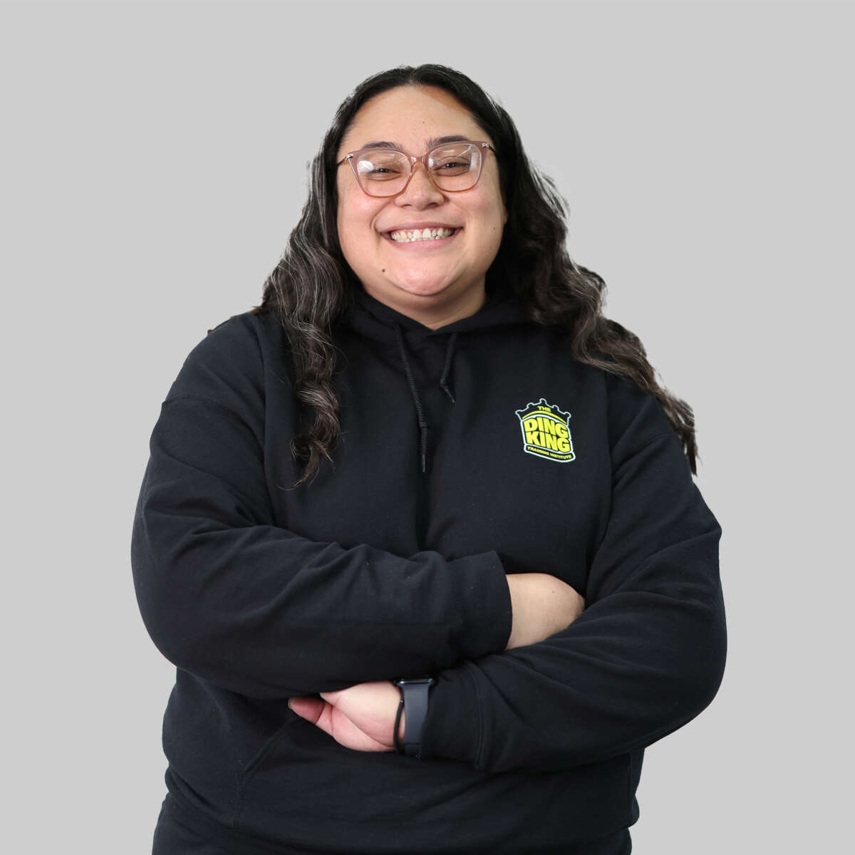 A woman in a black hoodie smiling with arms crossed, standing against a gray background for our "Meet the Team" staff profiles.