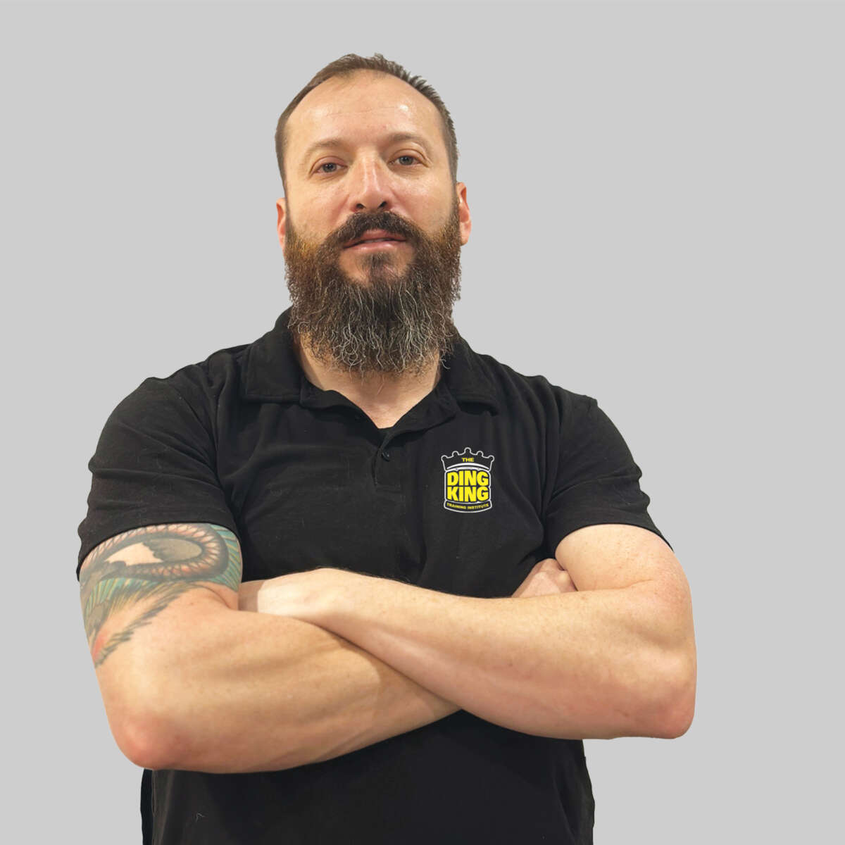 A man with a beard, wearing a black polo shirt with a "Ding King" logo, stands with arms crossed, displaying tattoos, against a gray background in our "Meet the Team" section