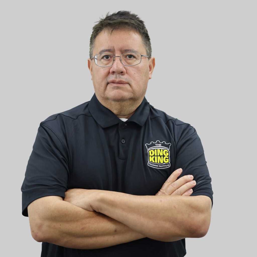 Meet the team: A man with short hair and glasses stands confidently with his arms crossed, wearing a black polo shirt featuring a logo that reads "The Ding King." The backdrop is a simple gray.