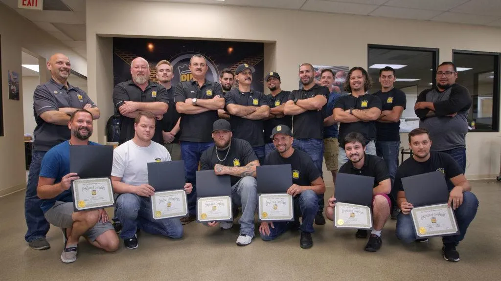 A group of men in a room, with some holding certificates from Advanced PDR Training and others standing behind them. The men holding certificates are kneeling or sitting, while the others pose with folded arms.