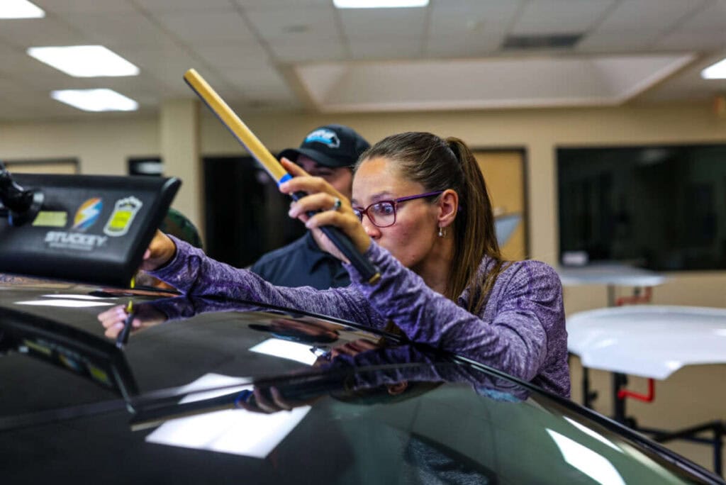 A person wearing glasses and a purple shirt utilizes advanced PDR training techniques to repair the roof of a car inside a well lit workshop.