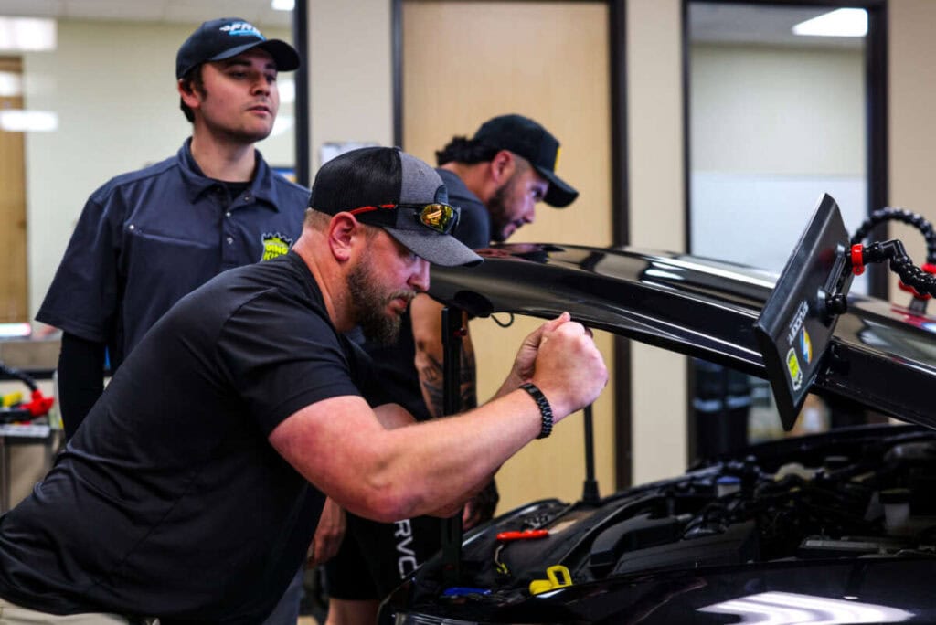 Three men work on a car's engine bay in a garage, focusing on repairs. One man wears a black shirt and cap, another in a navy shirt observes, while the third concentrates on the engine. They seem to be engaged in advanced PDR training to refine their skills.