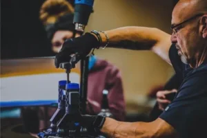 A person wearing black gloves operates machinery that seems to involve precise handling of components, showcasing their skills from advanced PDR training. Another individual in the background appears to be observing attentively.