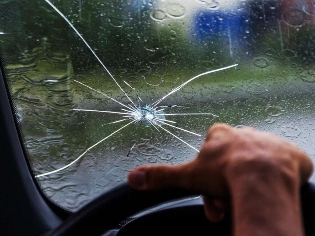 A cracked windshield of a car with raindrops visible outside. The driver's hand is gripping the steering wheel, reflecting their frustration from missing out on paint protection film training.