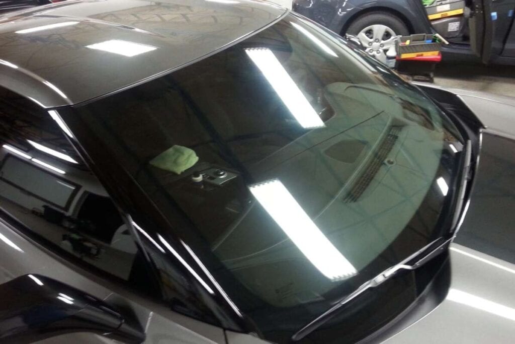 Close up view of a car windshield reflecting overhead garage lights. A white cloth is on the dashboard, and another vehicle is visible in the background with an open door, possibly involved in Paint Protection Film Training.