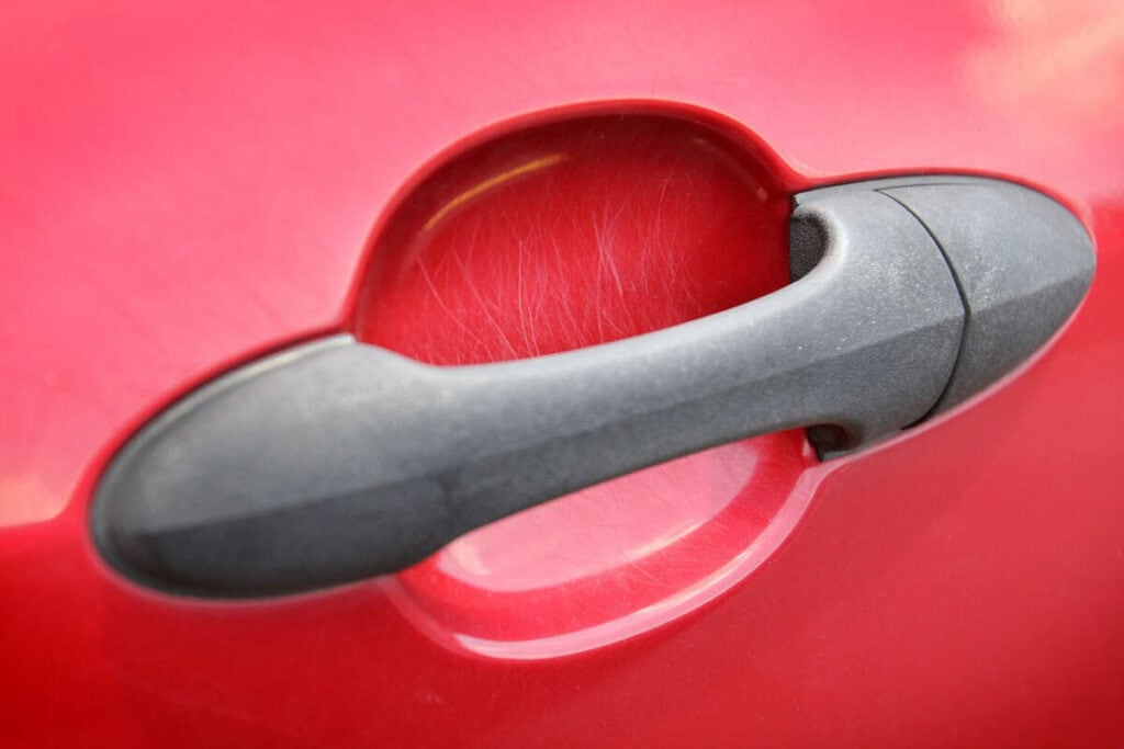 Close up of a red car door handle with minor scratches surrounding the handle area. The black handle is situated in the center of the image, highlighting the importance of Paint Protection Film Training to prevent such damage.