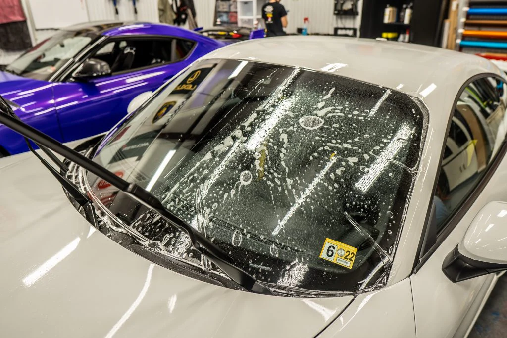 Two cars inside an auto detailing shop, one white and one blue. The white car has cleaning foam on its windshield with wipers raised. Nearby, technicians are engaged in Paint Protection Film Training to ensure every vehicle gets the best care possible.