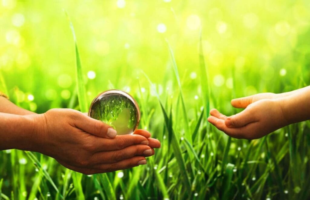 Two hands exchange a glass sphere over green grass and a bright, blurred background. The scene resembles the careful precision seen in paint protection film training, with the left hand appearing to be giving the sphere to the right hand.