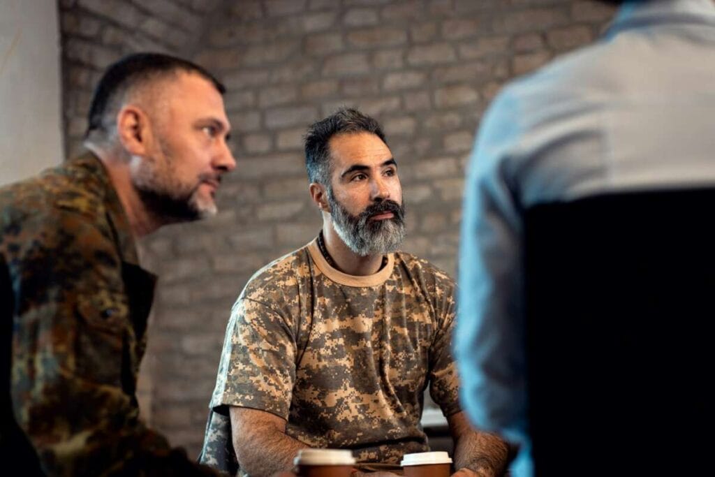 Three men, two in camouflage uniforms and one in civilian clothing, sit together at a table with two coffee cups in front of them against a brick wall background, discussing advanced PDR training.