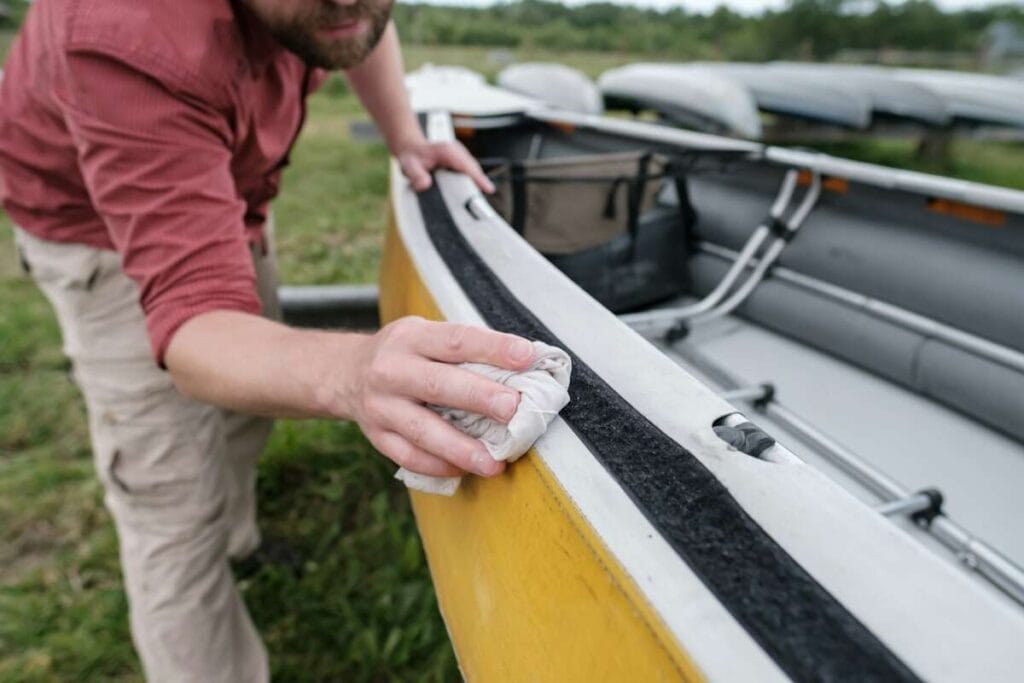 A person in a red shirt and beige pants is cleaning the edge of a yellow canoe with a cloth, with more canoes stacked in the background, possibly preparing for a Paint Protection Film Training.