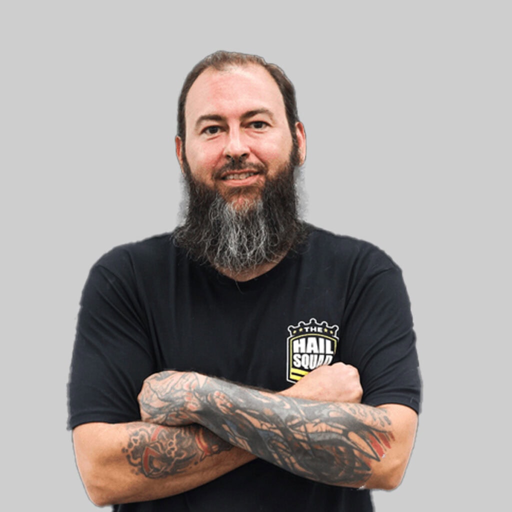 Meet the team: Man with a long beard and tattoos on his arms, wearing a black T shirt that has the "Hail Squad" logo, stands against a plain gray background with arms crossed.