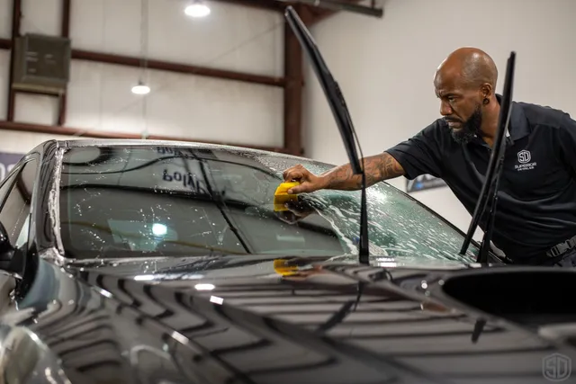 A man is cleaning the windshield of a black car with a sponge inside a garage, as part of his Paint Protection Film Training, with the windshield wipers raised.