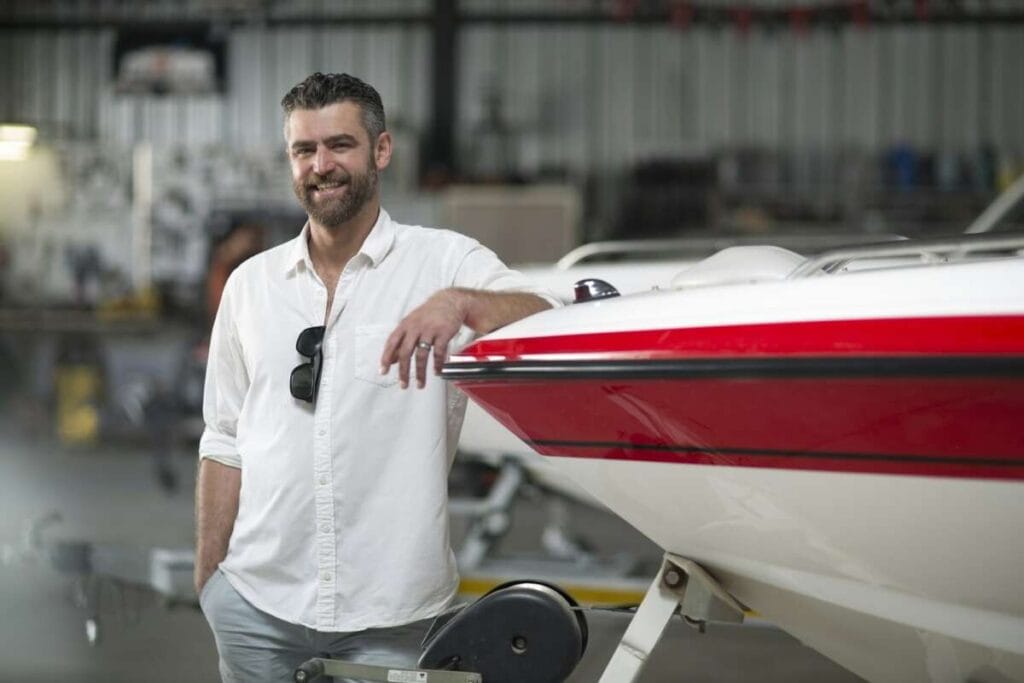 A man in a white shirt leans against a red and white boat in a well lit indoor setting, smiling at the camera after completing Paint Protection Film Training.