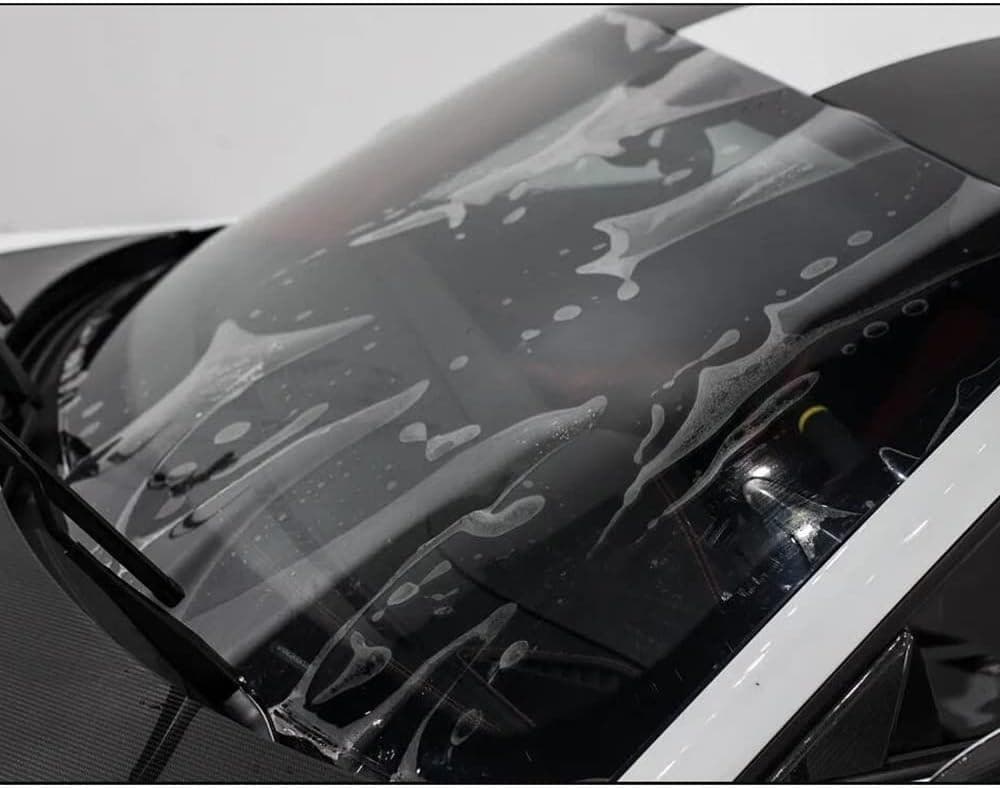 A close up of a car's windshield covered in soap and water, being cleaned. The windshield wipers are visible, and the reflection of the surroundings can be seen on the glass, indicative of a meticulous wash often performed during Paint Protection Film Training.