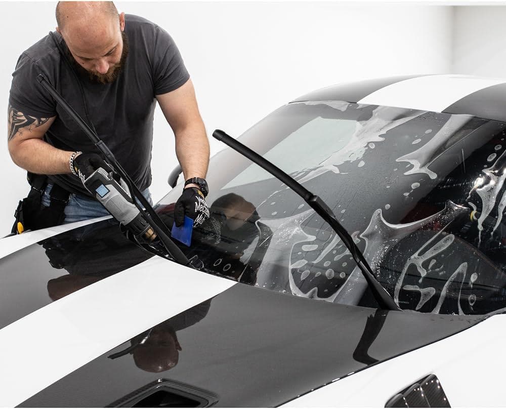 A man in a grey t shirt and gloves is applying a protective film to the black and white striped windshield of a car. With one hand holding a tool and the other a squeegee, he demonstrates techniques likely learned during Paint Protection Film Training.