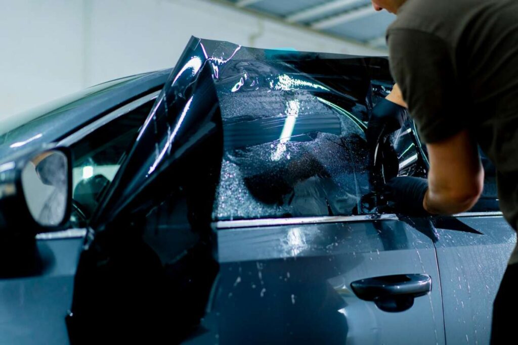 a car mechanic carefully sticks a protective tinted film on a car glass at a detailing station